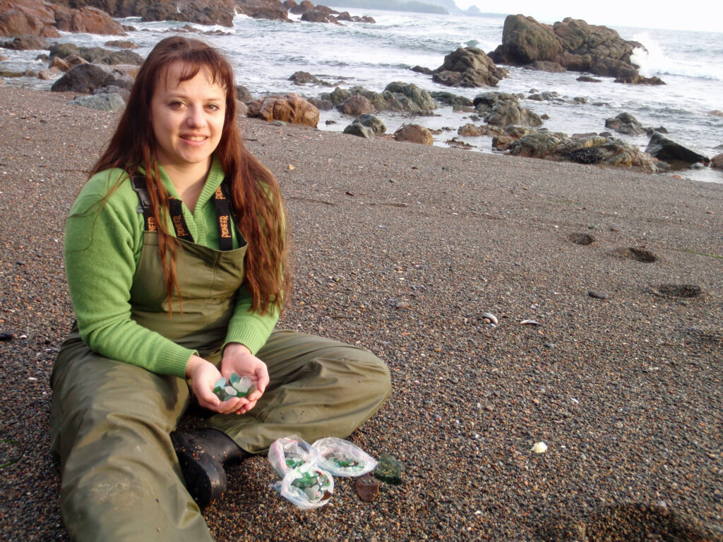 Chris sitting on a North Vancouver Island beach, holding a handful of green and aqua sea glass beside bags of the day’s finds